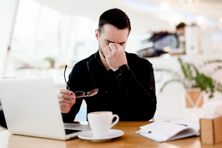 Give your eyes a rest Young tired freelancer man in black shirt suffers from pain and dryness in the eyes. He works a lot Cozy coffeehouse atmosphere on background. Cup of yummy cappuccino on table.の写真素材