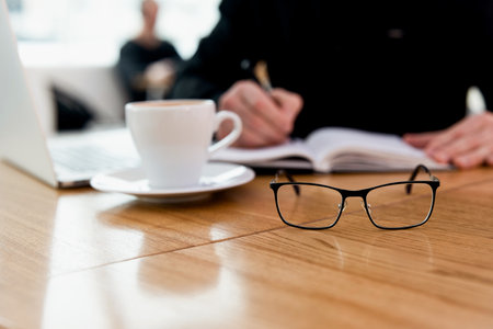 Man no longer needs glasses Young concentrated freelancer in black shirt writing a date of meeting in his diary and enjoying amazing cappuccino in coffee shop. Modern laprop and white mug on table.の写真素材
