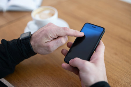 Man using his phone in coffee shop. Smartphone with blank copy space screen. Cup of cappuccino or flat white and open diary on wooden table. Mans hands hodling smartphone.の写真素材