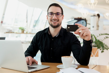 I can buy all I want Cheerful male remote worker showing his credit card, looking at camera and smiling. Happy man received a salary. He working from cafe, using his modern laptop.の写真素材