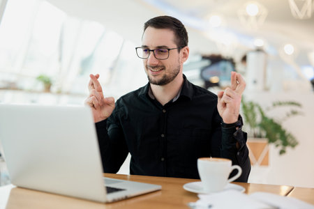 Man in eyeglasses and black shirt gesturing finger crossed and smiling. Luck and superstitious concept. Young attractive man with beard sitting in cafe, speaking by camera on his laptop.の写真素材