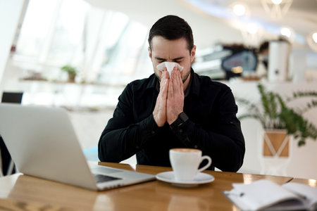 Sick man with runny nose came working in coffee shop. Tired man in black shirt blows his nose in public place. Isolate yourself if you get sick. Stay home concept. Cup of coffee and laptop on table.の写真素材