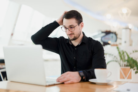 Hard remote work. Freelancer man holding his head doesnt understand what is happening in the chat with the employer. Male remote worker in black shirt working from coffeehouse and using laptop.の写真素材