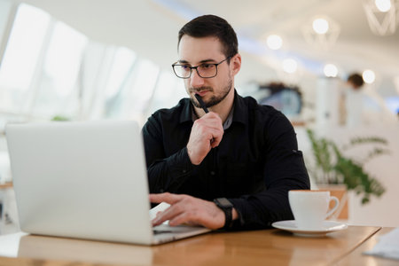 Concentrated man using laptop, holding pen near the mouth and thinking about his task. Work from cafe. Attractive intelligent man with beard wearing eyeglasses and black shirt programming.の写真素材