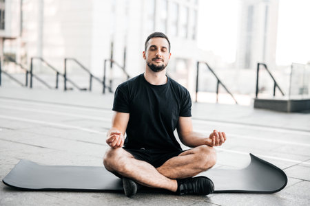 Man practicing yoga in big city. Sporty man meditating after workout. Excercising at home. Young man with stubble sitting on black yoga mat with eyes closed outdoors and relaxing in easy seat pose.の写真素材