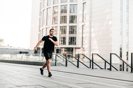 Young fit man doing urban running workout on asphalt road sprinting in sport and healthy lifestyle concept. Big city on background. Black sportswear. Caucasian man jogging.の写真素材
