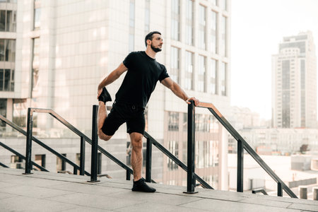 Athletic man stretching legs before workout outdoors. Runner in black sportswear excercising at...