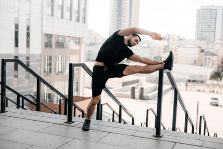 Strong man warming up. Morning activity. Man stretching his arms and looking away. Morning workout holding handrail. Black sportswear. Fit man doing tilts of the body to the side outdoors.の写真素材