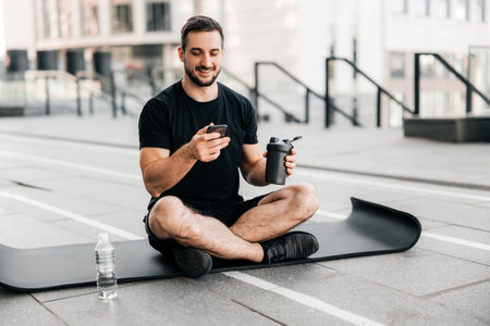 Man resting after outdoors workout with water and phone. Healthcare concept. Young sporty man after practicing yoga, break in doing exercise, relaxing on yoga mat, texting on smartphone.の写真素材