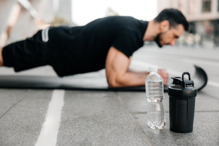 Concentrated man doing plank position while exercising on black yoga mat. Water bottles lying near. Blurred man in black sportswear exercising outdoors. Morning warm up. Side view.の写真素材