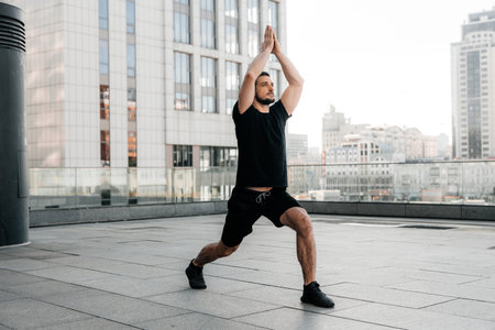 Athletic man practice Low Lunge pose with raised arms. Young man in black sports t-shirt and leggins stretching in yoga pose. Urban sport concept. Morning warm up.の写真素材