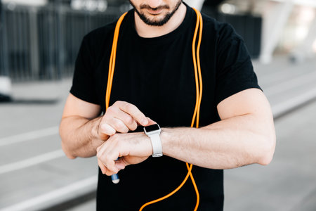 Athletic man with orange jumping rope around neck setting up his smart watch. Cut view of confitent male runner which records the results of the run on the device. Modern technologies concept.の写真素材