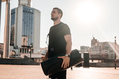 Confident young man holding rolled yoga mat and looking at beautiful morning view of big city. Morning activities concept. Bright sunshine on background. Black sporswear.の写真素材