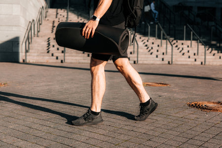 Tired and satisfied male sportsman returns home after morning workout. Cut view of sporty mans legs. Urban sport concept. Man walking on street and holding rolled yoga mat.の写真素材