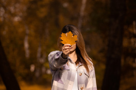 Women holding maple leaf. Fall is coming concept. Colorful autumn forest on background. Young woman wearing plaid blazer closed face with maple leaf in hand.の写真素材