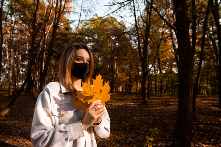 Woman in black medical face mask holding bright bouquet of yellow maple leaves. Walk during quarantine. Autumn foliage. Cozy fall season outdoors. Colorful autumn forest on background.の写真素材