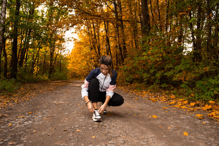 Young woman trail runner tying shoelaces in forest. Cozy fall season outdoors. Healthy lifestyle concept. Autumn foliage on road. Nice woman with ponytail wearing tracksuit jogging in park or forest.の写真素材