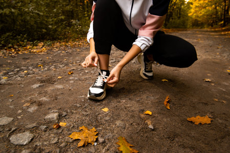 Womans hands fixing shoelaces on her sneakers. Close cut view. Female trail runner. Morning run concept. Young athletic woman jogging in forest. Healthy lifestyle concept. Autumn foliage on road.の写真素材