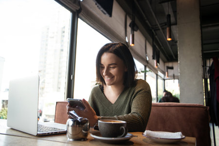 Happy young woman in cafe smiles looking into smartphone. Online communication concept. Pretty young woman sits in a coffee shop with laptop open before her and a cup of coffee.の写真素材