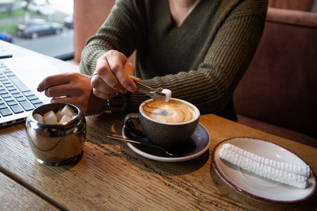 Woman in green pullover adds a piece of sugar to her cappucino with tongs while working at a coffee shop. Cozy cafe concept. Cut view of laptop on background.の写真素材