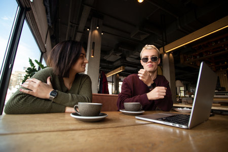 Couple working together in cafe. Young brunette woman smiling and looking at cool blonde man in black round glasses. Gray laptop on table. Two cups of coffee. Freelance working concept.の写真素材