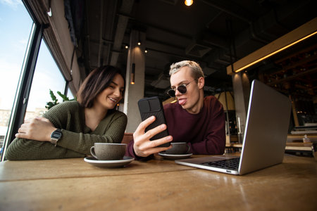 Man showing memes to woman. Lunch break concept. Young attractive man showing tongue and funny message on his smartphone to beautiful brunette woman. Working in coffee shop concept.の写真素材