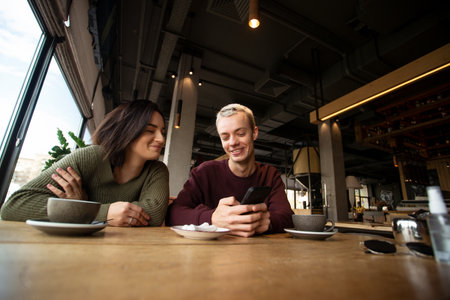 Young couple resting in a cafe and having fun. Man with blonde short hair showing something laughable in his phone barely holding back a smile. Cozy atmosphere in coffee shop or cafe.の写真素材