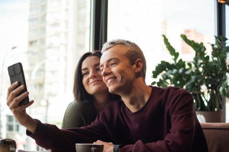 Happy couple taking selfie in cafe. Young attractive blonde man in purple sweatshort holding camera and taking photo with pretty brunette woman. Big light window on background. Restaurant or cafe.の写真素材