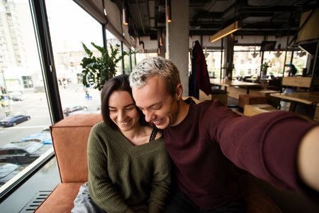 Selfie of young cheerful couple. Blonde man holding a camera and embracing happy woman. Restaurant or cafe on background. Cozy atmosphere. Smart style concept. Public place. Caucasian couple.の写真素材