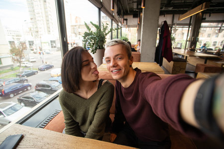 Couple makes crazy faces on camera. Coffee shop on background. Blond man with short hair holds camera and smiling when her girlfriend showing a tongue to him. Stylish man and woman.の写真素材