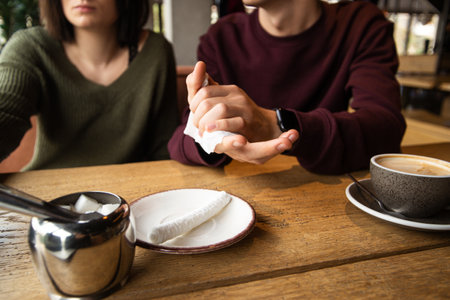 Man wipes his hands with a napkin. Caffe or restaurant on background. Man cleaning his hands by using white tissue paper. Cut view of young couple in public place. Cup of tasty coffee.の写真素材