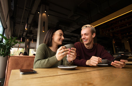 Caucasian couple drinks coffee in cafe. Brunette woman warms her hands on the cup she holds in her hands and smiles to blonde man in purple sweatshirt. Cozy atmosphere in coffee shop.の写真素材