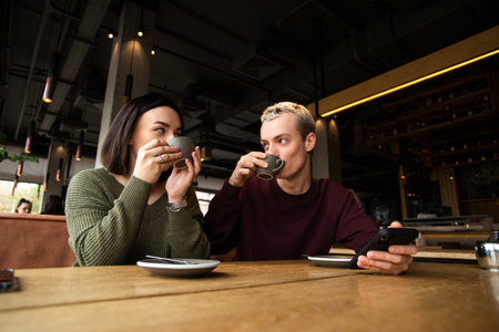 Young couple enjoys delicious coffee in cafe. They holding cappuccino cups and looking at each other. Tasty drinks. Coffee shop or restaurant on background.の写真素材
