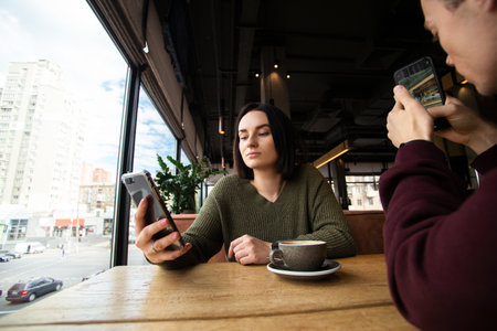 Young couple spends time on the phone while meeting each other. Social media concept. Brunette woman chatting in her smartphone and man taking photo of cafe. Modern communication concept.の写真素材