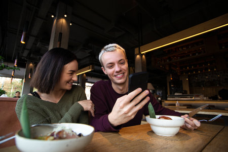 Couple eating in restaurant and laughing. Man showing memes to woman. Good mood concept. Young man showing something funny on his smartphone to beautiful brunette woman. Good relationship concept.の写真素材