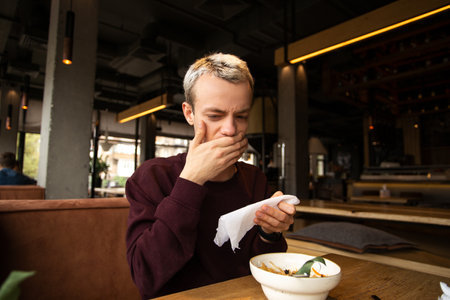 Spoiled food in the restaurant. Dissatisfied young man in a cafe covers his mouth with a hand disgusted with a taste of food. A meal on the table in front of him. Bad customer service concept.の写真素材