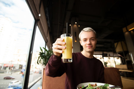 Good looking young man sits in a cafe near panoramic window. He holds a glass of orange juice in his hand and shows it to camera. Caesar salad. Pleasant meal in cafe.の写真素材