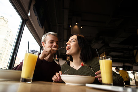 Man feeding his girlfriend a tomato. WAttractive woman smiling and dreamly smiling. She is happy that she will get more delicious salad. Two glasses of orange fresh on table. Healthy diet on cafe.の写真素材