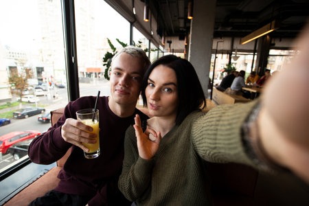 Young couple posing for selfie in public place. Attractive brunette woman and good looking blond man take photo in a restaurant together. Man holds a glass of orange juice. Woman makes okay gesture.の写真素材