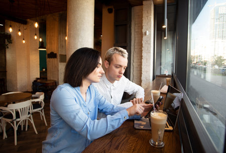 Two freelance workers in coffee house. Brunette woman showing something in her smartphone and pointing on screen, man listening. Decision making concept. Cup of latte and gray laptop on table.の写真素材