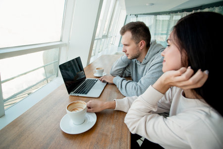 Young couple spending time in cafe and wathcing video on laptop. Woman and man drinking coffee and looking photos togehter. Cafe on background. Woman fixing her hair when man using laptop.の写真素材