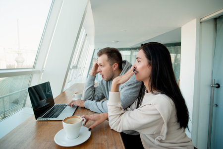 Couple saw something shoking on laptop. Man fiddles with hair and woman giggles and covers her mouth with hand. Empty coffee cup. White light room on background.の写真素材