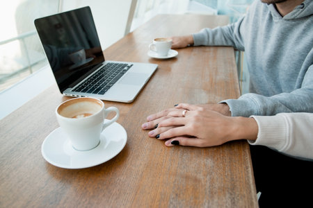 Womans hand with gold ring covers mans hand. Romantic date concept. Modern laptop and two empty coffee cups on wooden table. Casual style. Heterosexual couple good spending time.の写真素材