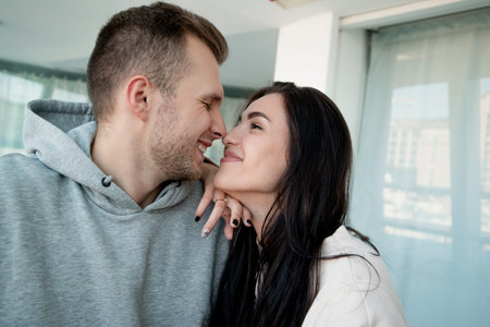 Beautiful couple in love embraced and gently rubs noses. Brunette woman with long hair looking deeply into mans eyes. White light room on background. Young cheerful couple touched one anothers nose.の写真素材