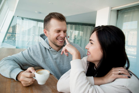 Cheerful couple having a good time with a cup of coffee. Happy dark haired woman pointin by her...