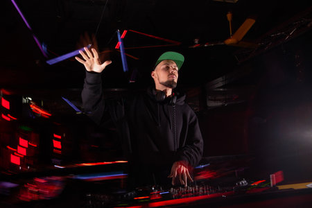 Charismatic disk jockey enterntains crowd at techno party. Feel the rhythm. Blurred red and blue stage lights on background. Smart nightclub interior. Satisfied DJ plays top tracks.の写真素材