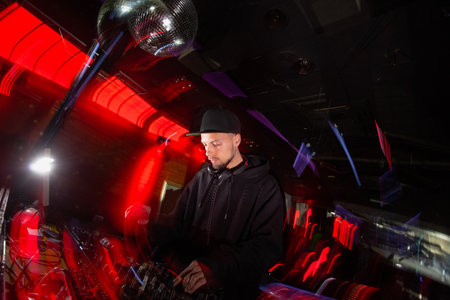Concentrated DJ plays music on a party. Good looking young man in black hat and casual black clothes uses turntable to mix music. Blurred background with red light.の写真素材