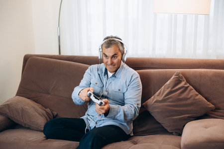 Adult man playing on console. Good time on weekend concept. Concentrated man with headphones sitting on brown sofa, holding game controller and playing video games.の写真素材