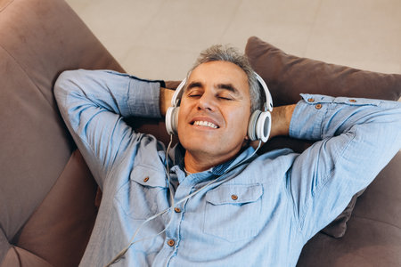 Middle aged man listening to music, lying on sofa with closed eyes and smiling with teeth. Casual style. Man enjoying his favorite music and relaxing at home. Spending time during quarantine.の写真素材