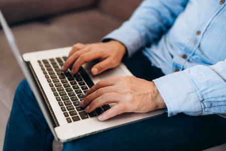 Close up male hands typing at keyboard. Middle aged man using modern laptop to read news or reply to emails. Blue shirt and jeans. Modern technology concept.の写真素材
